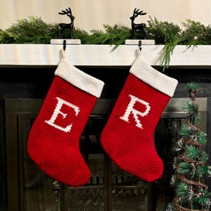 May include: Two red Christmas stockings with white cuffs and large white letters "E" and "R". The stockings hang from black deer-shaped hooks on a white mantel, decorated with greenery.