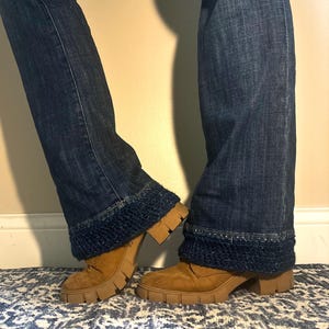 May include: A pair of blue denim jeans with a dark blue knit cuff detail. The jeans are paired with brown suede boots with a chunky, lug sole. The boots have a low heel and a rounded toe. The jeans are flared at the bottom.