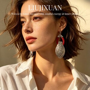 以下が含まれることがあります： 赤い織りのアクセントと詳細な花の模様が施されたシルバー色のイヤリング。 イヤリングは涙の形をしており、シルバーのフックから吊り下げられています。 画像には「LIUJIXUAN」と「Woven by hand, blessed with soulful energy & inner peace」というテキストが含まれています。
