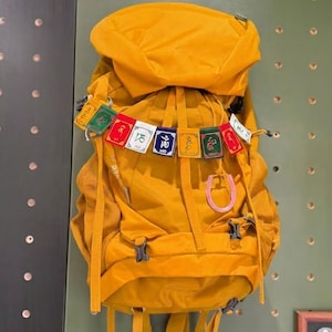 May include: Mustard yellow backpack with multiple compartments and straps. A string of colourful prayer flags hangs from the top. The backpack is hanging on a green wall with small, evenly spaced holes.