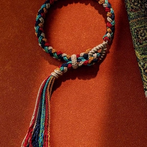 Puede incluir: Una pulsera de macramé hecha a mano con un diseño trenzado colorido. La pulsera presenta una mezcla de hilos rojos, azules, amarillos y blancos, con una borla en el extremo. La pulsera se muestra sobre un fondo naranja.