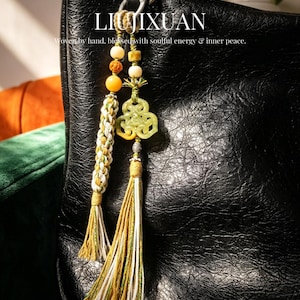 Puede incluir: Un bolso de mano de cuero negro con un adorno decorativo. El adorno presenta un cordón trenzado, cuentas amarillas y crema, y un colgante color jade. El texto "LIUJIXUAN" y un lema son visibles.