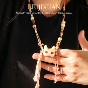 May include: A necklace featuring a central cream-colored fox mask pendant, accented with brown, cream, and metallic beads on a woven cord. The piece includes pink and yellow tassels and displays the text LIUJIXUAN Woven by hand, blessed with soulful energy & inner peace.