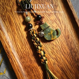 Puede incluir: Un llavero artesanal con un dije en forma de gato verde, un cordón trenzado negro y verde y cuentas decorativas. El llavero incluye el texto "LIUJIXUAN" y la frase "Woven by hand, blessed with soulful energy & inner peace."