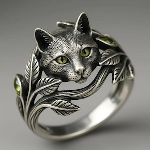 May include: A silver ring featuring a detailed cat head with bright green eyes, surrounded by sculpted metal leaves and vines. The band is crafted from polished silver, incorporating organic leaf motifs that wrap around the finger.