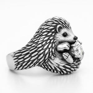 May include: A silver hedgehog ring with detailed quills and a small face. The hedgehog is holding a clear, faceted gemstone. The ring has a smooth band and is set against a white background.
