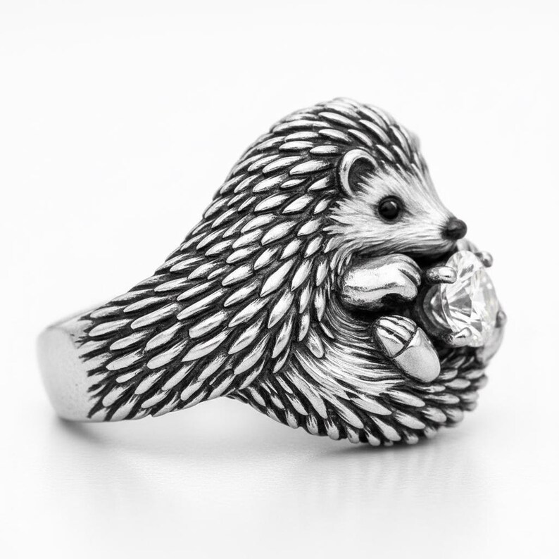 May include: A silver hedgehog ring with detailed quills and a small face. The hedgehog is holding a clear, faceted gemstone. The ring has a smooth band and is set against a white background.