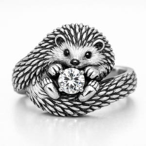 May include: A silver-colored ring featuring a detailed hedgehog design. The hedgehog is holding a large, clear, round gemstone. The ring has a textured band and the hedgehog's quills are intricately detailed.