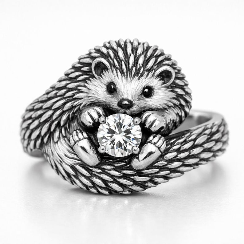 May include: A silver-colored ring featuring a detailed hedgehog design. The hedgehog is holding a large, clear, round gemstone. The ring has a textured band and the hedgehog's quills are intricately detailed.