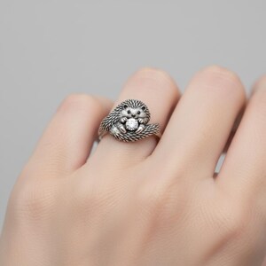 May include: A silver hedgehog ring with a detailed design. The hedgehog is holding a clear, round gemstone. The ring is worn on a finger, showcasing the intricate details of the jewelry.