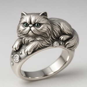 May include: A silver-toned ring featuring a detailed sculpture of a Persian cat with green eyes. The cat is lying on the ring band, which is embellished with small, clear gemstones. The ring has a vintage or antique aesthetic.