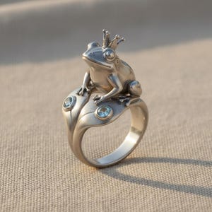 Könnte beinhalten: Ein silberfarbener Ring mit einem skulptierten Frosch, der eine kleine Krone trägt und auf zwei stilisierten Blättern sitzt. Jedes Blatt ist mit einem einzelnen, runden, hellblauen Edelstein verziert, der in die Metalloberfläche eingelassen ist.