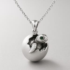 Może przedstawiać: Srebrny naszyjnik z wisiorkiem przedstawiającym małego żółwia morskiego wychodzącego z pękniętego jaja. Żółw ma zielone oko z kamienia szlachetnego. Wisiorek zawieszony jest na delikatnym łańcuszku, biżuteria inspirowana naturą.