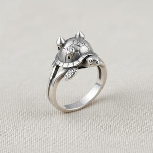 Puede incluir: Un anillo de tono plateado con un diseño de tortuga donde el caparazón tiene forma de pequeña torre de castillo. El caparazón de la tortuga está decorado con dos pequeñas piedras redondas y transparentes incrustadas en el metal, con una banda lisa.