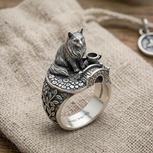 May include: A silver ring featuring a sculpted cat sitting on a textured surface next to a small cup and a miniature arched bridge. The band is decorated with floral patterns and the interior is engraved with the word HANDCRAFTED.