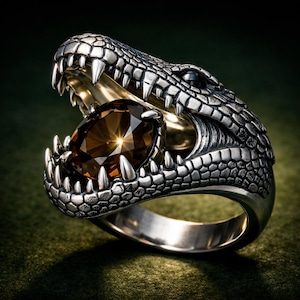 May include: A silver-colored ring featuring a detailed crocodile head design. The crocodile's open mouth holds a large, brown gemstone. The ring has a textured, scale-like pattern and sharp teeth.