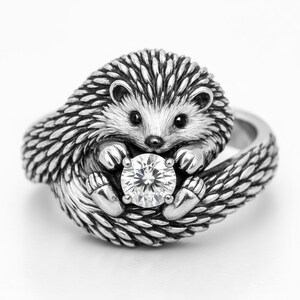 May include: A silver-toned ring featuring a detailed hedgehog design. The hedgehog is curled around a large, clear, round gemstone. The ring's design includes textured quills and paws, creating a whimsical and detailed piece of jewelry.