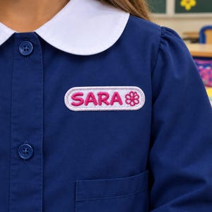 Pode incluir: Uma camisa azul-escura de manga comprida com gola branca arredondada e fecho de botões. No peito, há um emblema retangular branco bordado com o nome SARA em letras cor-de-rosa ao lado de um pequeno ícone de uma flor rosa.