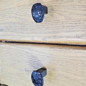 May include: Close-up of two dark blue, oval-shaped drawer pulls with a textured design. The pulls are mounted on a light-colored wooden drawer. The drawer pulls have a rectangular base.