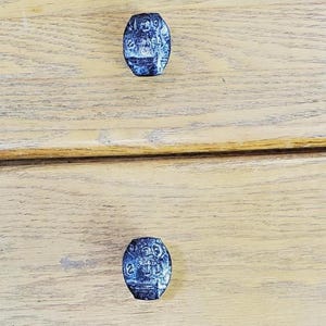 May include: Two oval-shaped cabinet knobs with a dark blue, textured surface are mounted on a light-colored wooden drawer. The knobs have a detailed, possibly engraved design.