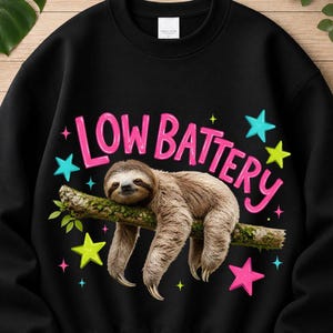 May include: A black crewneck sweatshirt featuring a graphic of a brown sloth resting on a tree branch. Above the sloth, the text "LOW BATTERY" is printed in pink letters. The design is accented with colorful stars in shades of pink, blue, and yellow.