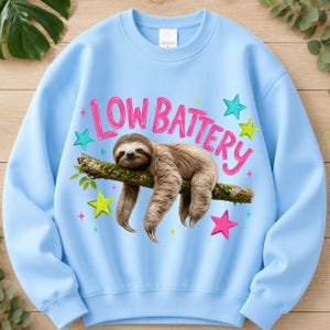 Puede incluir: Una sudadera de cuello redondo en color azul claro con un gr&aacute;fico de un perezoso marr&oacute;n colgado de una rama. Sobre el perezoso, el texto "LOW BATTERY" est&aacute; impreso en color rosa brillante. El dise&ntilde;o incluye varias estrellas coloridas en tonos rosa, amarillo y turquesa.