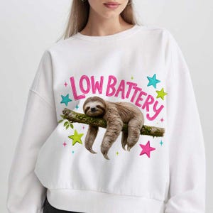 Puede incluir: Una sudadera blanca de cuello redondo con un gr&aacute;fico de un perezoso marr&oacute;n colgado de una rama. Sobre el perezoso, el texto "LOW BATTERY" est&aacute; impreso en rosa. El dise&ntilde;o est&aacute; decorado con estrellas coloridas en tonos azul, verde y rosa.