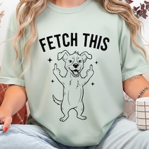 Może przedstawiać: Jasnozielona koszulka z napisem "FETCH THIS" nad czarno-białą ilustracją psa z kreskówki. Pies pokazuje środkowy palec obiema rękami. Cztery czarne gwiazdy otaczają psa.