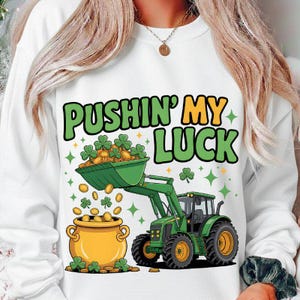 Op de afbeelding: Witte sweatshirt met een St. Patrick's Day-ontwerp. Het toont een groene tractor die gouden munten en klavers in een pot schept. De tekst "PUSHIN' MY LUCK" is in groen en geel.