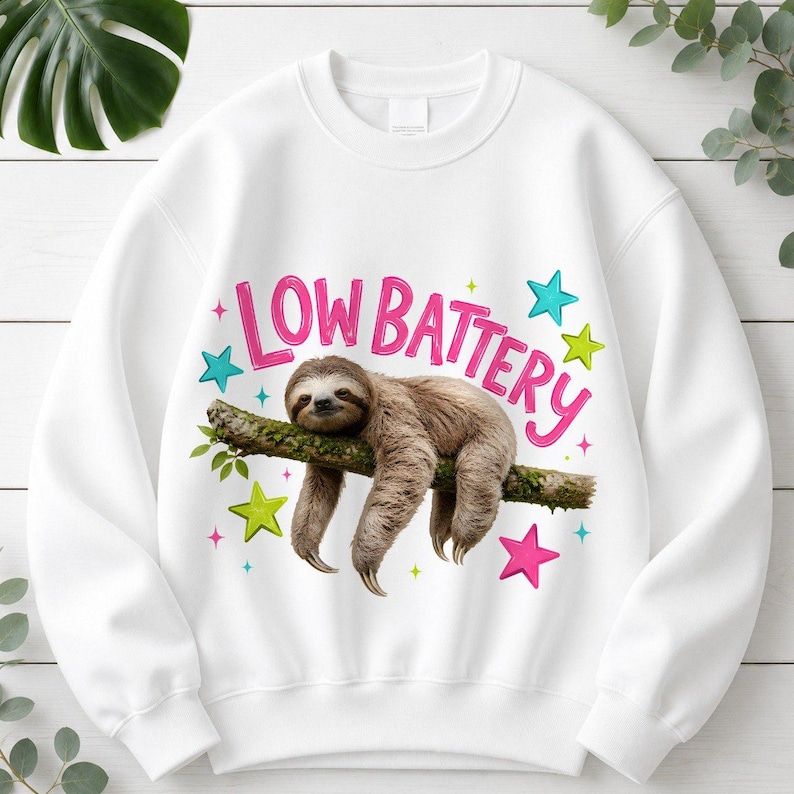 Puede incluir: Una sudadera blanca de cuello redondo con un gr&aacute;fico de un perezoso marr&oacute;n colgado de una rama de &aacute;rbol. Sobre el perezoso, el texto "LOW BATTERY" est&aacute; impreso en letras rosas. El dise&ntilde;o est&aacute; decorado con estrellas coloridas en azul, verde y rosa.