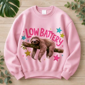 Puede incluir: Una sudadera de cuello redondo rosa claro con un gr&aacute;fico de un perezoso marr&oacute;n colgado de una rama. Sobre el perezoso, el texto LOW BATTERY aparece en letras rosas. El dise&ntilde;o est&aacute; decorado con estrellas coloridas en tonos azul, rosa y amarillo.