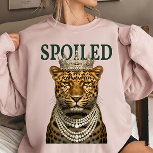 Op de afbeelding: Een lichtroze sweatshirt met een luipaard met een kroon en een parelketting. Het woord "SPOILED" is in donkergroen boven de kop van de luipaard gedrukt. De luipaard heeft een gevlekte vacht.