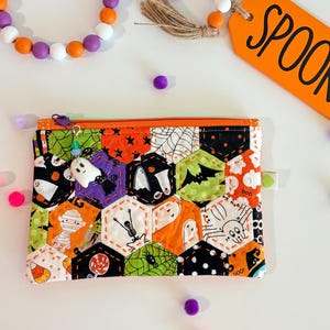 May include: A wooden bead garland with orange, purple, and white beads, jute tassels, and an orange tag with the word SPOOKY in black text, paired with a rectangular zippered pouch featuring a quilted hexagonal pattern of Halloween motifs.