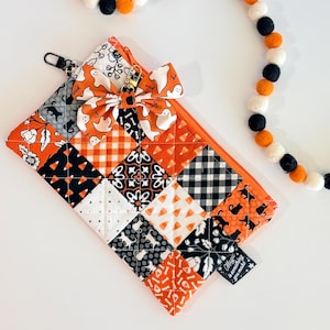 May include: A quilted zipper pouch featuring a patchwork pattern of orange, black, and white fabrics with ghost, cat, and geometric prints. A fabric bow is attached to the zipper pull, accompanied by a long garland of felt balls in orange, black, and white.