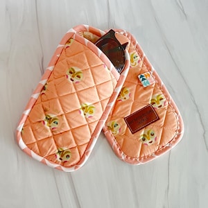 Może przedstawiać: Pikowane etui na okulary przeciwsłoneczne w kolorze brzoskwiniowym z powtarzającym się wzorem twarzy zwierzęcia z kreskówki. Etui ma wzór w romby i jest obszyte biało-różową tkaniną w paski. W środku znajdują się okulary przeciwsłoneczne.
