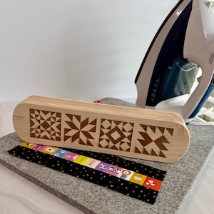 Op de afbeelding: Een lichtgekleurde houten kist met een geometrisch ontwerp, naast een zwarte stoffen strook met kleurrijke tekst en illustraties. Een grijs strijkplankje en een blauw-wit strijkijzer zijn ook zichtbaar.