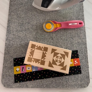 May include: A gray felt ironing mat with a wooden block featuring a geometric design, a pink rotary cutter, and a white iron. A black fabric with colorful squares and stars is also present.