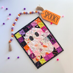 Könnte beinhalten: Ein rechteckiger, gesteppter Wandbehang mit einem zentralen Geistermotiv, umgeben von einem Patchwork-Rand aus orangefarbenen, violetten, schwarzen und weißen Stoffquadraten. Eine Holzperlengirlande in Orange, Violett und Weiß ist befestigt und endet an einem Schild mit der Aufschrift SPOOKY.