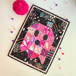 May include: A rectangular quilted wall hanging featuring a central skull motif composed of various pink and purple patterned fabrics. The border is black with white polka dots and a pink trim. It includes a label reading Wolf's Bane.