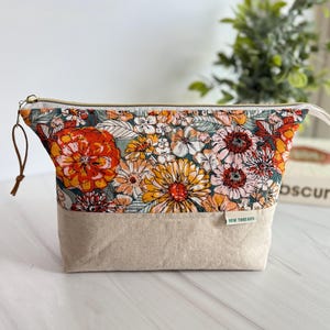 May include: A floral print cosmetic bag with a beige bottom and a gold zipper. The bag features a vibrant pattern of orange, yellow, and pink flowers on a green background. The tag reads "NEW THREADS".