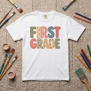 Puede incluir: Una camiseta blanca de manga corta con el texto FIRST GRADE en letras grandes de estilo bloque. Cada letra tiene un patrón único, incluyendo verde oliva sólido, cuadros vichy rojos y blancos, estampados florales y beige, en dos filas.