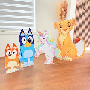 Pode incluir: Recortes de cartão de personagens de desenhos animados, incluindo um cão azul, um cão laranja, um unicórnio com uma crina arco-íris e uma leoa. Os personagens estão em pé em um chão de madeira, com uma janela e elementos decorativos no fundo.