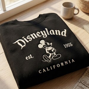 Op de afbeelding: Zwart sweatshirt met het woord "Disneyland" in witte letters, een Mickey Mouse-afbeelding en de tekst "est. 1955 CALIFORNIA". Het sweatshirt is op een houten oppervlak gevouwen.