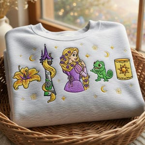 May include: White sweatshirt featuring embroidered images of a yellow flower, a tower, a princess, a green chameleon, and a lantern. The sweatshirt is in a woven basket.