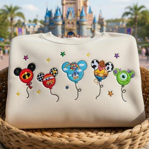 Puede incluir: Sudadera color crema con bordados de globos de personajes de dibujos animados. Los globos son Mickey Mouse, Rayo McQueen, un globo azul de Mickey con nubes, un vaquero y Mike Wazowski. La sudadera est&aacute; en una cesta de mimbre.
