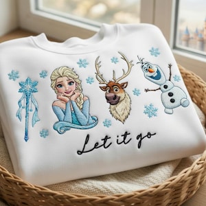 以下が含まれることがあります： 白のスウェットシャツ。刺繍されたエルサ、オラフ、スヴェン、雪の結晶、雪の結晶の杖が特徴です。「Let it go」の文字が刺繍されています。スウェットシャツは柳のバスケットに折りたたまれています。