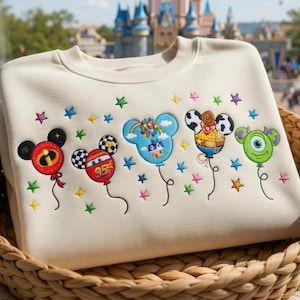 Puede incluir: Sudadera de color crema con globos de personajes de dibujos animados bordados. Los globos incluyen Mickey Mouse, un coche de carreras, un arco&iacute;ris, Woody y Mike Wazowski. Estrellas de colores est&aacute;n bordadas encima de los globos. La sudadera est&aacute; doblada en una cesta tejida.