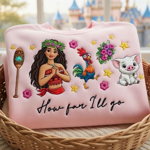 Może przedstawiać: Jasnor&oacute;żowa bluza z haftowanymi postaciami z kresk&oacute;wek, w tym kobietą, kogutem i świnią, a także elementami kwiatowymi. Tekst "How far I'll go" jest wyhaftowany na dole. Bluza jest prezentowana w wiklinowym koszu.