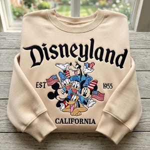 May include: Beige sweatshirt with the word "Disneyland" in black script. Below the word are cartoon characters including Mickey Mouse, Minnie Mouse, Donald Duck, and Goofy. The sweatshirt also has the words "EST 1955" and "CALIFORNIA".