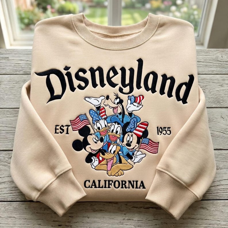 May include: Beige sweatshirt with the word "Disneyland" in black script. Below the word are cartoon characters including Mickey Mouse, Minnie Mouse, Donald Duck, and Goofy. The sweatshirt also has the words "EST 1955" and "CALIFORNIA".